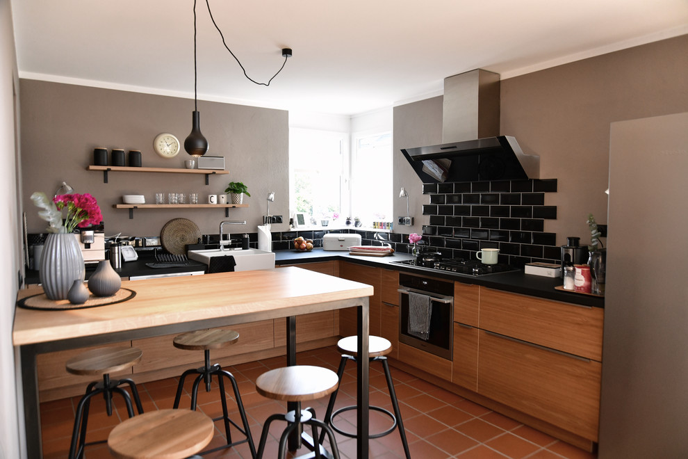 Design ideas for a mid-sized industrial l-shaped open plan kitchen in Frankfurt with a single-bowl sink, flat-panel cabinets, medium wood cabinets, wood benchtops, black splashback, subway tile splashback, stainless steel appliances, terra-cotta floors, a peninsula, red floor and black benchtop.