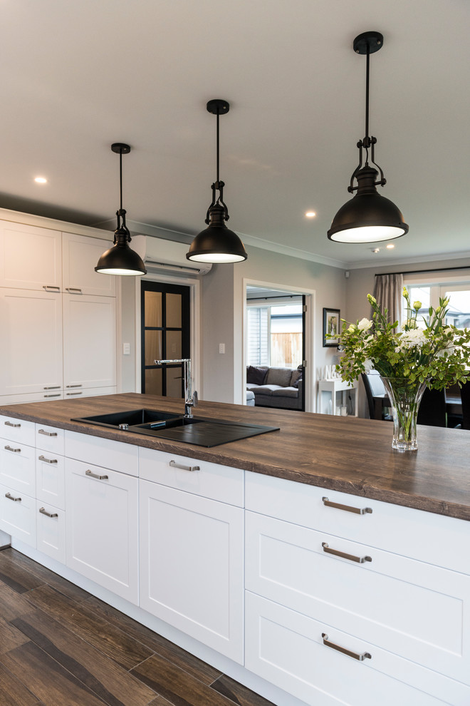 Inspiration for a large transitional l-shaped kitchen pantry in Christchurch with a farmhouse sink, shaker cabinets, white cabinets, laminate benchtops, black splashback, glass sheet splashback, stainless steel appliances, vinyl floors, with island and brown floor.