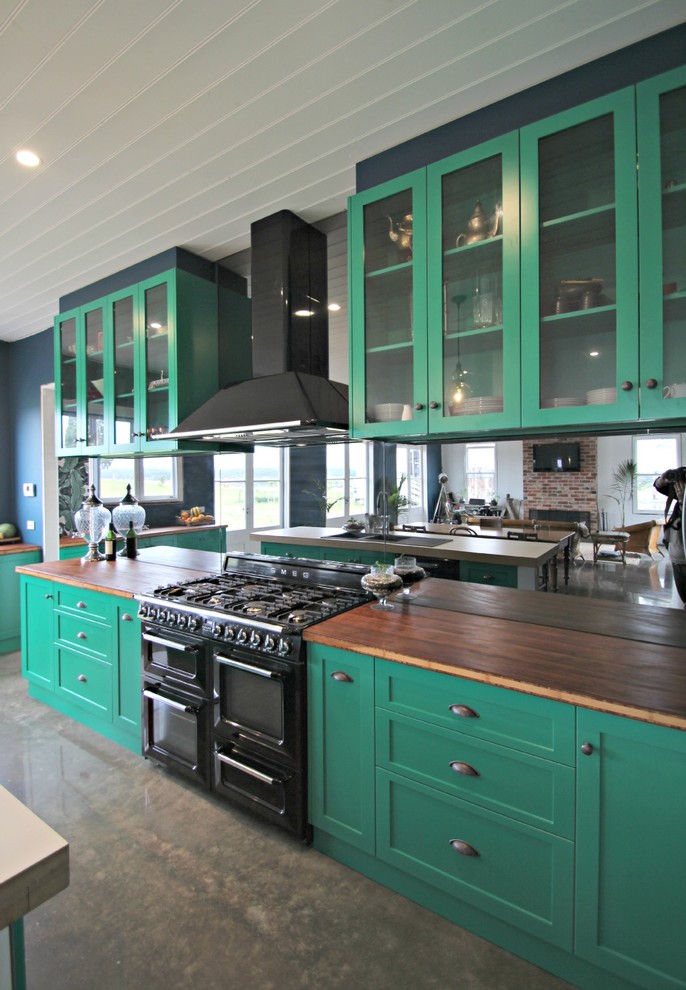 This is an example of a large eclectic l-shaped eat-in kitchen in Melbourne with a farmhouse sink, shaker cabinets, green cabinets, wood benchtops, metallic splashback, mirror splashback, black appliances, concrete floors and with island.