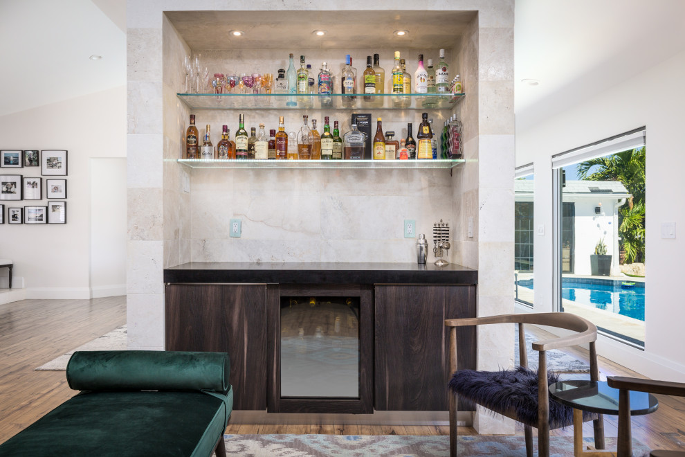 This is an example of a small contemporary single-wall dry bar in Miami with flat-panel cabinets, dark wood cabinets, engineered stone countertops, beige splashback, marble splashback, light hardwood flooring and black worktops.