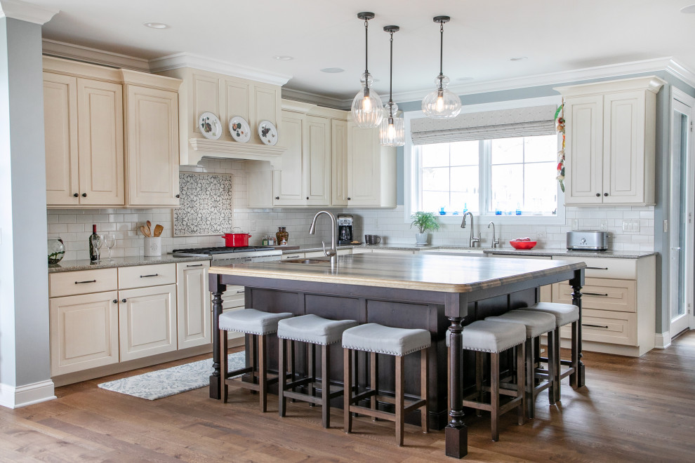Photo of a large traditional l-shaped kitchen in Milwaukee with a farmhouse sink, raised-panel cabinets, stainless steel appliances, medium hardwood floors, with island, beige cabinets, white splashback, subway tile splashback, brown floor, grey benchtop and granite benchtops.