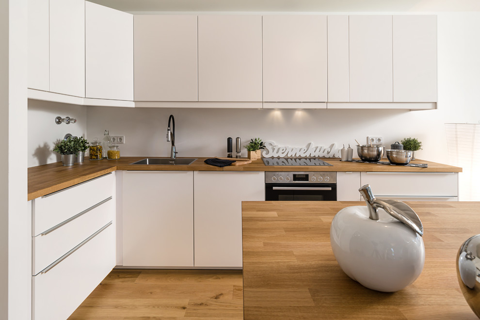 Small contemporary l-shaped open plan kitchen in Munich with medium hardwood floors, brown floor, a drop-in sink, flat-panel cabinets, white cabinets, wood benchtops, white splashback, black appliances, with island and brown benchtop.