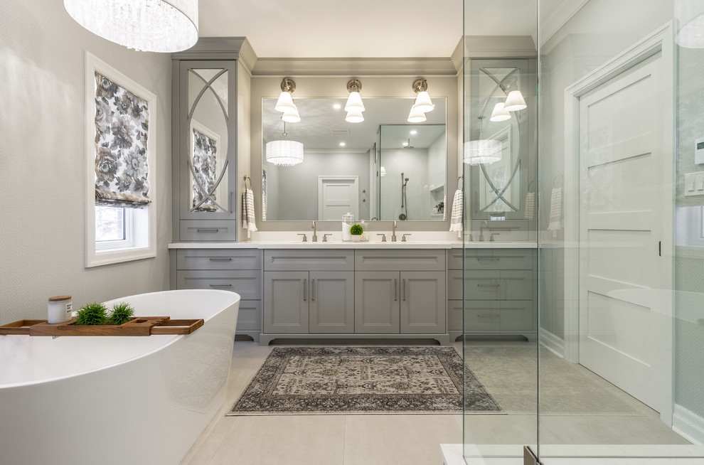 This is an example of a transitional master bathroom in Toronto with recessed-panel cabinets, grey cabinets, a freestanding tub, grey walls, an undermount sink, a hinged shower door, white benchtops, a corner shower and grey floor.