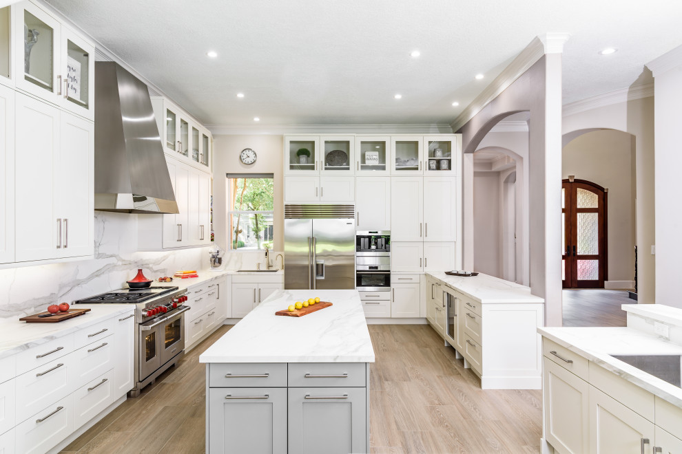 This is an example of a large transitional l-shaped eat-in kitchen in Houston with an undermount sink, shaker cabinets, white cabinets, marble benchtops, white splashback, marble splashback, stainless steel appliances, light hardwood floors, multiple islands, brown floor and white benchtop.
