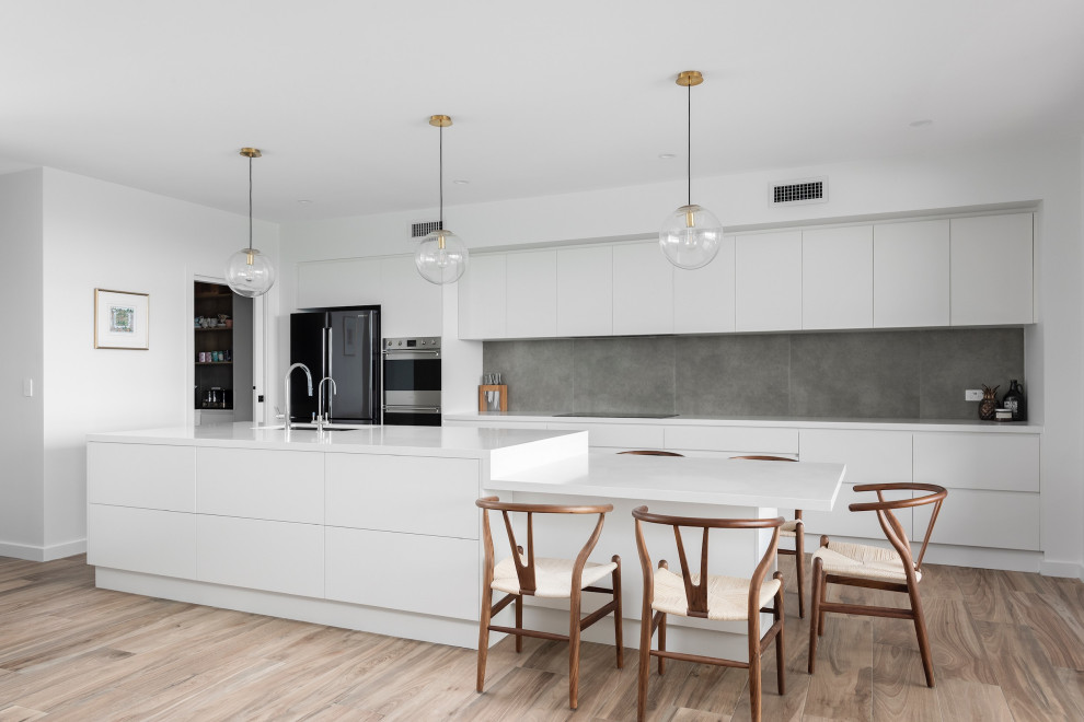 This is an example of a modern galley kitchen in Wollongong with an undermount sink, flat-panel cabinets, white cabinets, grey splashback, stainless steel appliances, light hardwood floors, with island, beige floor and white benchtop.