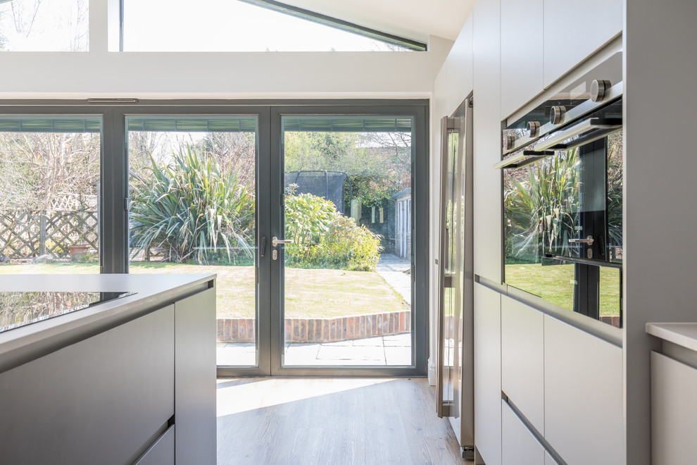 This is an example of a mid-sized contemporary galley open plan kitchen in Sussex with an integrated sink, flat-panel cabinets, grey cabinets, solid surface benchtops, stainless steel appliances, with island and white benchtop.