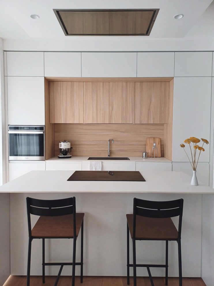 Photo of a medium sized scandi galley open plan kitchen in Milan with a submerged sink, flat-panel cabinets, white cabinets, composite countertops, wood splashback, stainless steel appliances, light hardwood flooring, an island and white worktops.