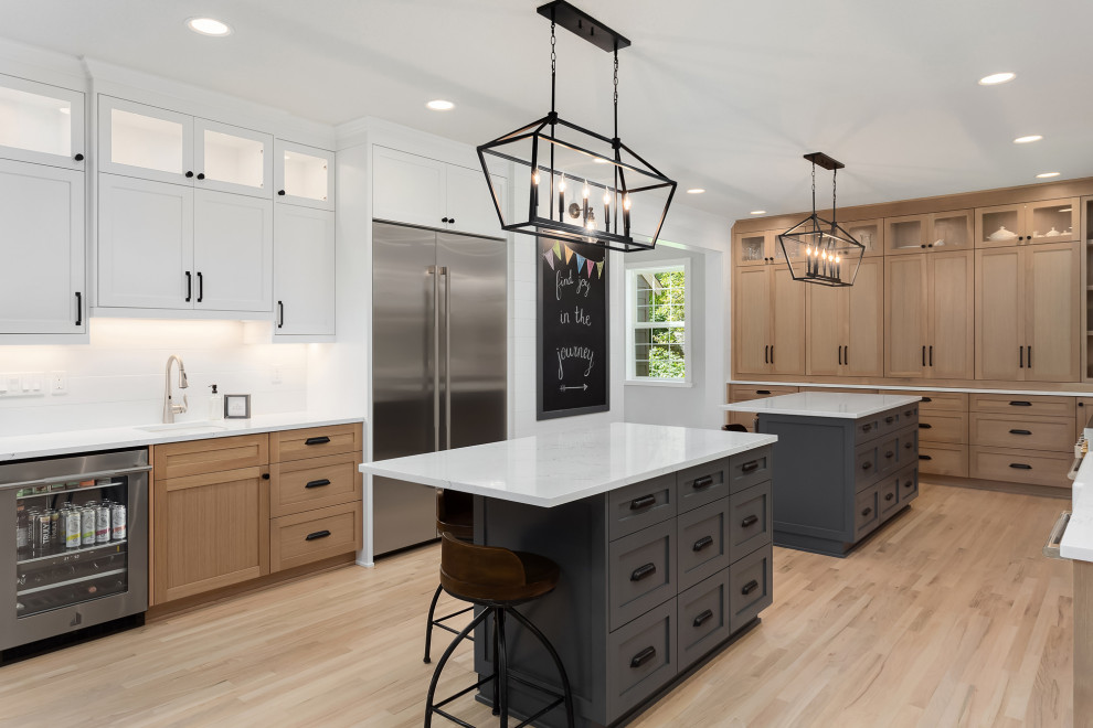 Large country l-shaped eat-in kitchen in Portland with a farmhouse sink, shaker cabinets, light wood cabinets, quartz benchtops, white splashback, cement tile splashback, stainless steel appliances, light hardwood floors, multiple islands and white benchtop.