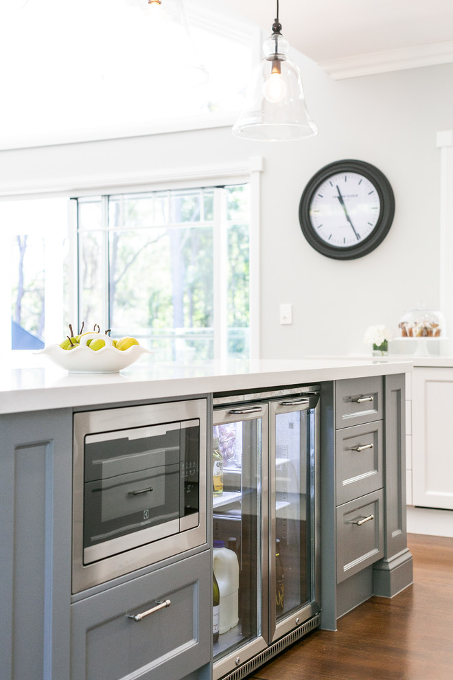 Design ideas for a large country kitchen in Sunshine Coast with a farmhouse sink, recessed-panel cabinets, white cabinets, quartz benchtops, white splashback, subway tile splashback, stainless steel appliances, dark hardwood floors, with island, brown floor and white benchtop.