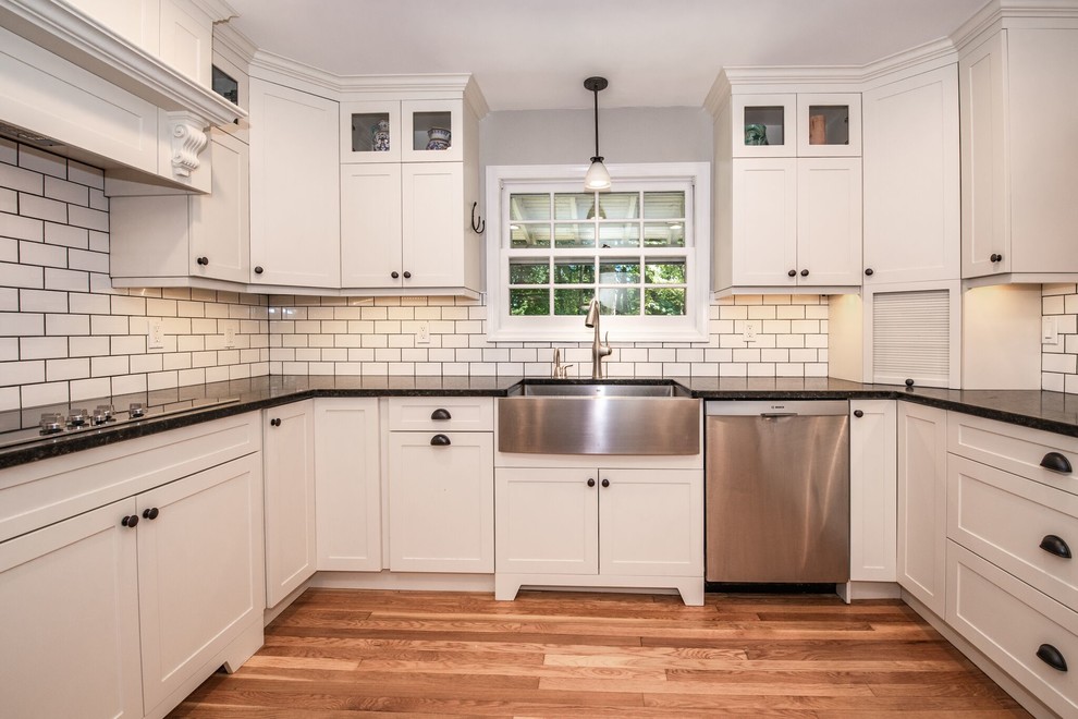 Large transitional u-shaped eat-in kitchen in New York with a farmhouse sink, shaker cabinets, white cabinets, solid surface benchtops, white splashback, subway tile splashback, stainless steel appliances, medium hardwood floors, a peninsula, brown floor and black benchtop.