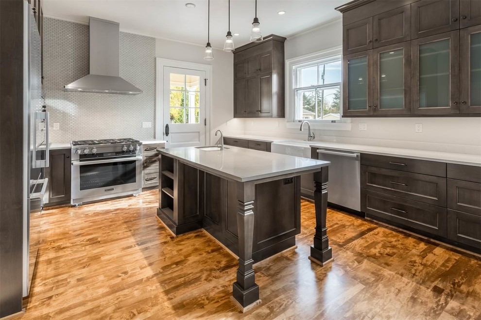 Inspiration for a mid-sized transitional u-shaped medium tone wood floor enclosed kitchen remodel in Seattle with a farmhouse sink, recessed-panel cabinets, gray cabinets, quartz countertops, white backsplash, ceramic backsplash, stainless steel appliances and an island