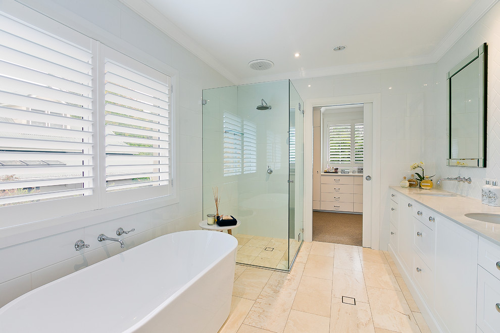 Inspiration for a mid-sized transitional master bathroom in Sydney with recessed-panel cabinets, white cabinets, a freestanding tub, a corner shower, white tile, ceramic tile, white walls, travertine floors, an undermount sink and a hinged shower door.