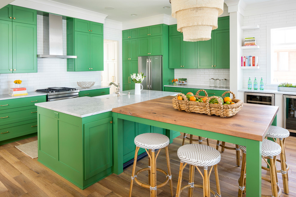 Inspiration for a large beach style l-shaped open plan kitchen in Minneapolis with a farmhouse sink, shaker cabinets, green cabinets, white splashback, subway tile splashback, stainless steel appliances, light hardwood floors, multiple islands, brown floor and marble benchtops.