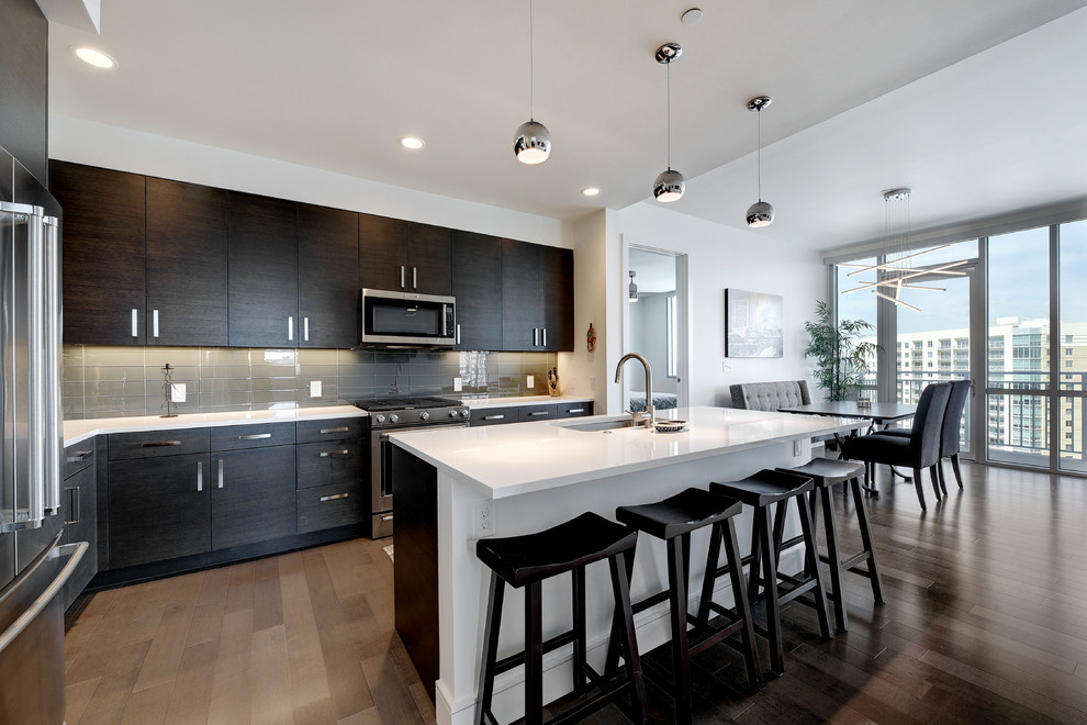 Photo of a contemporary l-shaped open plan kitchen in Austin with an undermount sink, flat-panel cabinets, dark wood cabinets, grey splashback, glass tile splashback, stainless steel appliances, dark hardwood floors, with island and white benchtop.