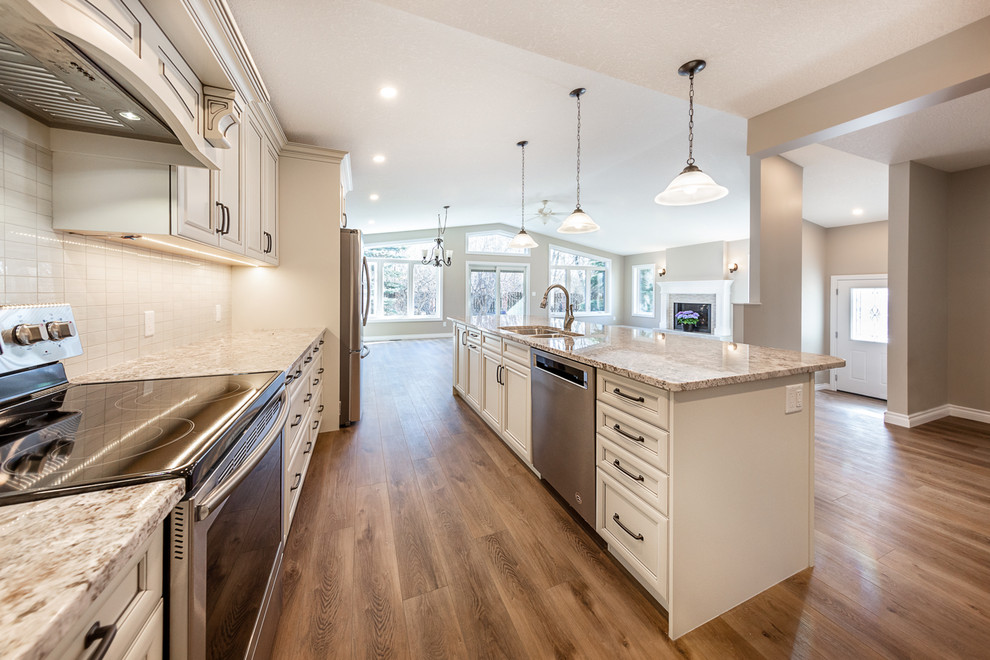 Photo of a large traditional l-shaped open plan kitchen in Edmonton with an undermount sink, shaker cabinets, white cabinets, granite benchtops, white splashback, ceramic splashback, stainless steel appliances, medium hardwood floors, with island, brown floor and beige benchtop.