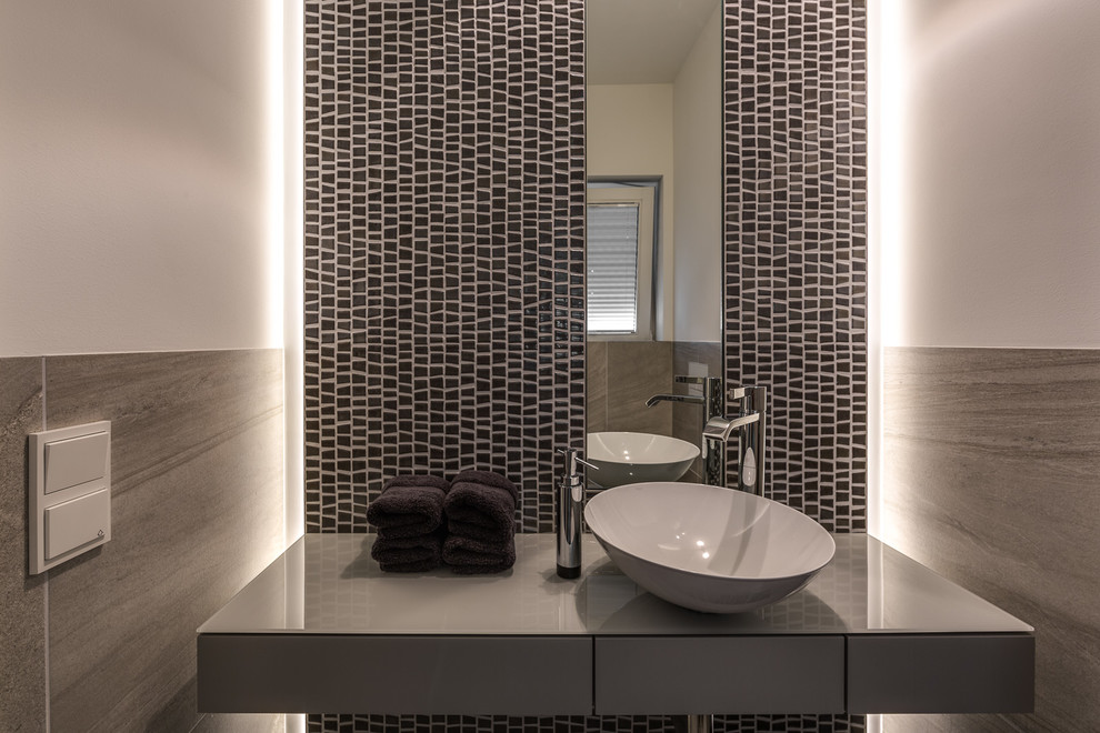 Inspiration for a small contemporary powder room in Cologne with flat-panel cabinets, grey cabinets, gray tile, mosaic tile, white walls, a vessel sink and glass benchtops.