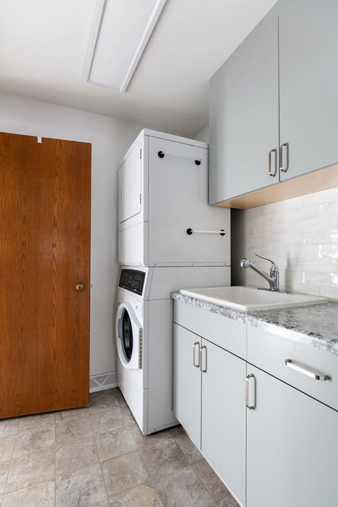 Photo of a small transitional single-wall dedicated laundry room in Other with a drop-in sink, flat-panel cabinets, grey cabinets, solid surface benchtops, white splashback, white walls, vinyl floors, a stacked washer and dryer, multi-coloured floor, multi-coloured benchtop and porcelain splashback.