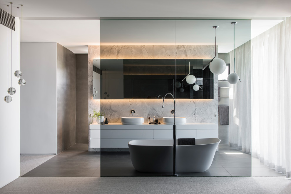 Photo of a large modern master bathroom in Sydney with furniture-like cabinets, white cabinets, a freestanding tub, an open shower, a wall-mount toilet, gray tile, marble, grey walls, porcelain floors, a vessel sink, solid surface benchtops, grey floor, an open shower and white benchtops.