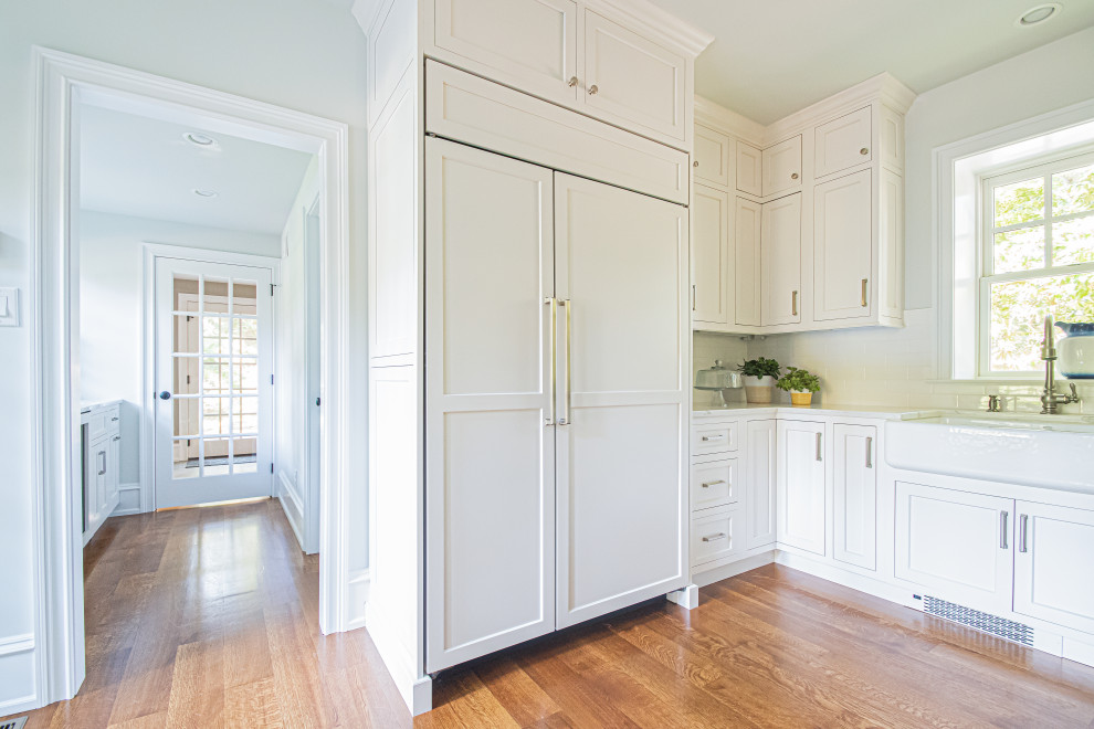 Inspiration for a large traditional l-shaped eat-in kitchen in Philadelphia with a farmhouse sink, recessed-panel cabinets, white cabinets, quartzite benchtops, white splashback, marble splashback, panelled appliances, medium hardwood floors, with island, brown floor and white benchtop.