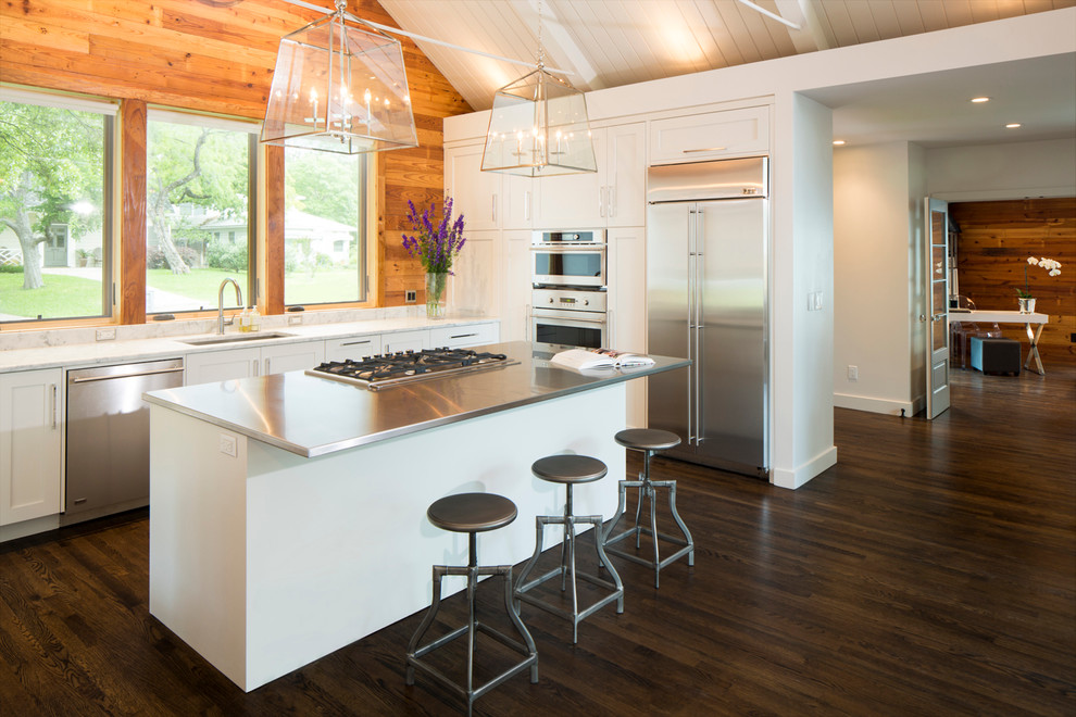Transitional u-shaped open plan kitchen in Austin with white cabinets, stainless steel appliances, dark hardwood floors, with island, shaker cabinets, stainless steel benchtops, white splashback, an undermount sink and brown floor.