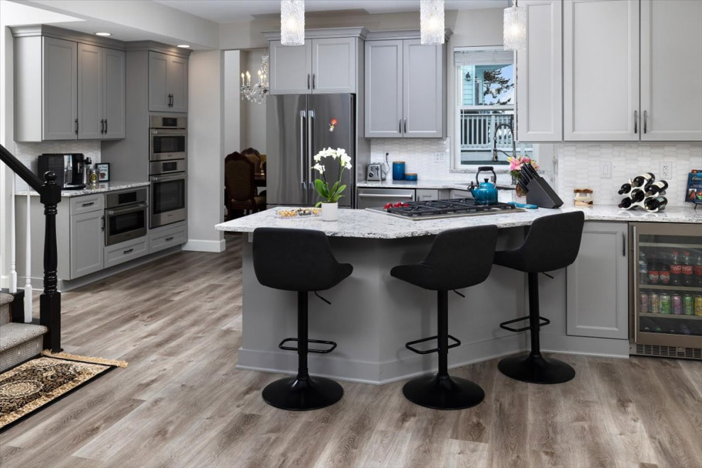 Example of a mid-sized transitional laminate floor and gray floor open concept kitchen design with recessed-panel cabinets, gray cabinets, quartz countertops, white backsplash, marble backsplash, stainless steel appliances and white countertops