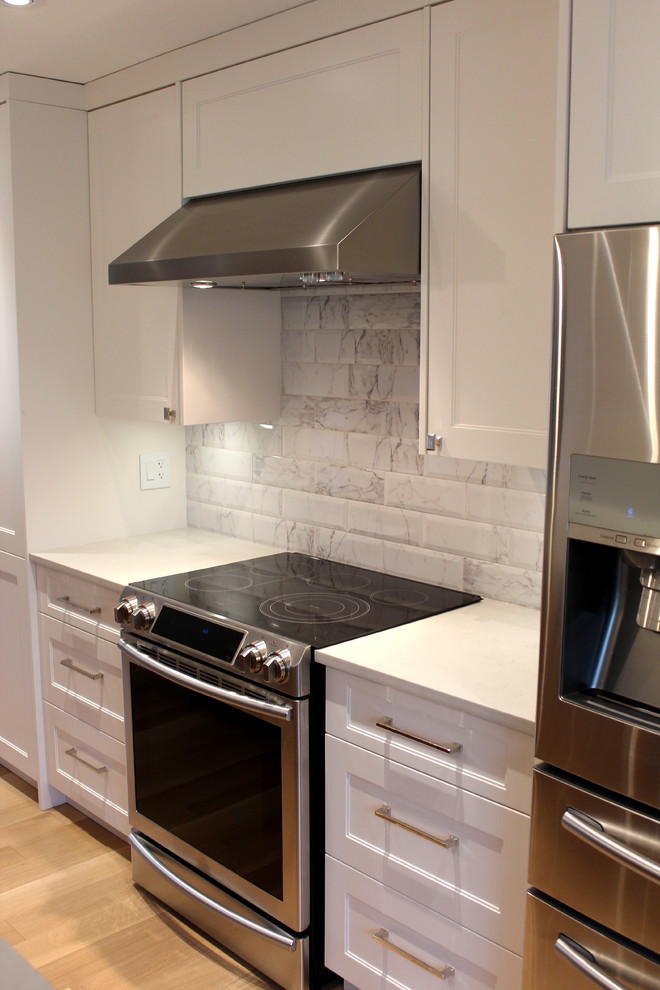 Large transitional l-shaped light wood floor eat-in kitchen photo with an undermount sink, shaker cabinets, white cabinets, quartz countertops, white backsplash, ceramic backsplash, stainless steel appliances and an island