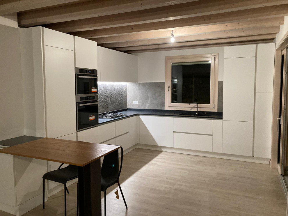 Inspiration for a mid-sized modern l-shaped eat-in kitchen in Venice with an integrated sink, white cabinets, quartz benchtops, black splashback, slate splashback, stainless steel appliances, laminate floors, a peninsula, black benchtop and exposed beam.