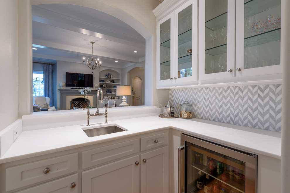 This is an example of a mid-sized traditional l-shaped wet bar in Dallas with an undermount sink, glass-front cabinets, white cabinets, granite benchtops, multi-coloured splashback, ceramic splashback, dark hardwood floors, brown floor and white benchtop.
