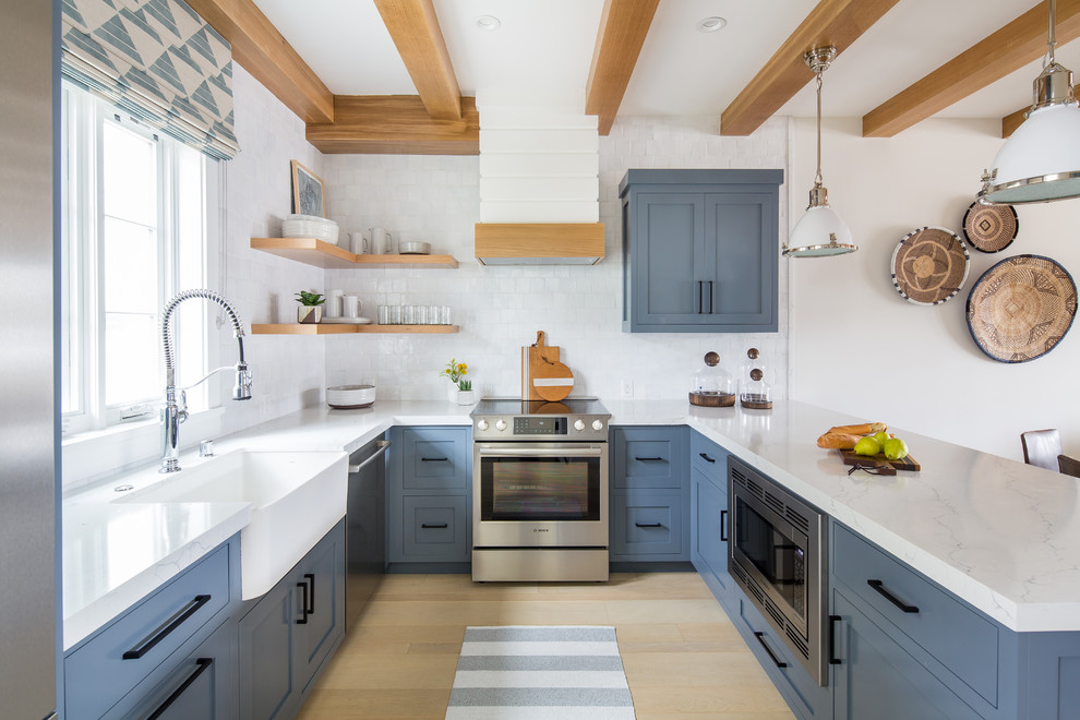 Photo of a beach style u-shaped kitchen in Orange County with a farmhouse sink, shaker cabinets, blue cabinets, white splashback, stainless steel appliances, light hardwood floors, a peninsula, beige floor and white benchtop.