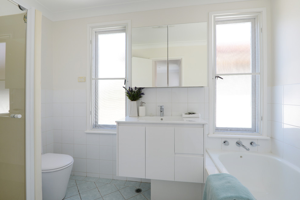 This is an example of a mid-sized contemporary master bathroom in Sydney with an integrated sink, white cabinets, an alcove tub, an alcove shower, a one-piece toilet, white tile, white walls and ceramic floors.