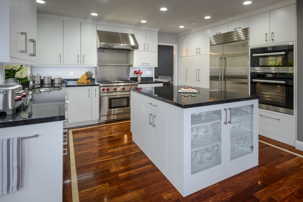 This is an example of a large modern l-shaped eat-in kitchen in Los Angeles with a drop-in sink, flat-panel cabinets, white cabinets, stainless steel appliances, medium hardwood floors, with island, black splashback, granite benchtops and slate splashback.
