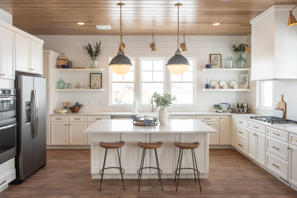 Design ideas for a beach style l-shaped kitchen in Salt Lake City with a farmhouse sink, recessed-panel cabinets, white cabinets, white splashback, timber splashback, stainless steel appliances, medium hardwood floors, with island and white benchtop.