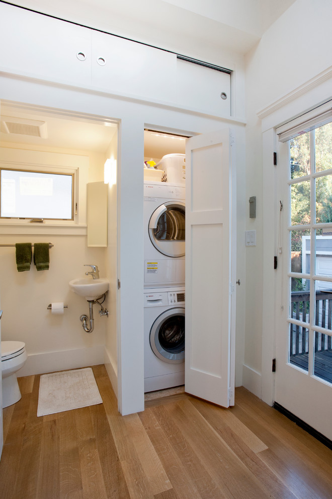 Photo of a small arts and crafts single-wall laundry cupboard in San Francisco with a single-bowl sink, shaker cabinets, medium hardwood floors, a stacked washer and dryer, white cabinets, white walls and brown floor.
