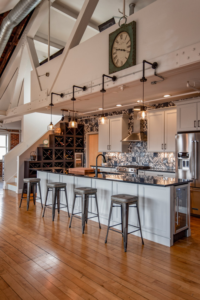 This is an example of a large industrial open plan kitchen in Providence with a single-bowl sink, recessed-panel cabinets, white cabinets, quartz benchtops, ceramic splashback, stainless steel appliances, light hardwood floors, with island and exposed beam.
