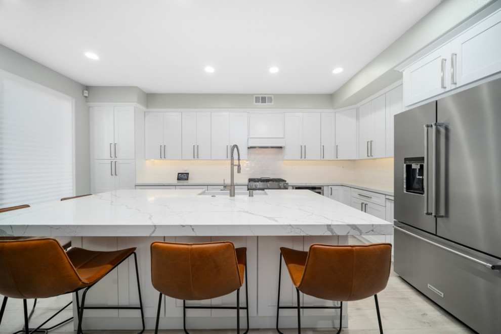 Mid-sized transitional laminate floor and gray floor kitchen photo in Orange County with a farmhouse sink, shaker cabinets, white cabinets, white backsplash, subway tile backsplash, stainless steel appliances, an island and white countertops