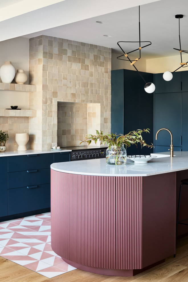 Inspiration for a large contemporary l-shaped kitchen/diner in London with a built-in sink, flat-panel cabinets, blue cabinets, composite countertops, beige splashback, ceramic splashback, black appliances, cement flooring, an island, pink floors, grey worktops and a chimney breast.