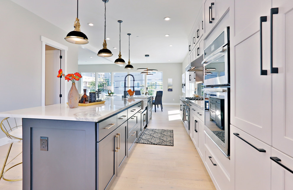 This is an example of a large contemporary galley open plan kitchen in Seattle with a farmhouse sink, shaker cabinets, white cabinets, granite benchtops, black splashback, marble splashback, stainless steel appliances, light hardwood floors, with island and white benchtop.