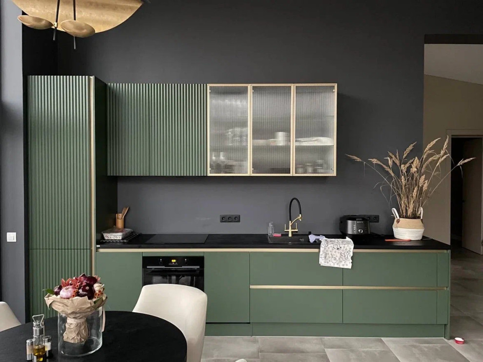 Design ideas for a mid-sized contemporary single-wall open plan kitchen in Moscow with a drop-in sink, flat-panel cabinets, green cabinets, solid surface benchtops, black splashback, black appliances, porcelain floors, no island, beige floor and black benchtop.