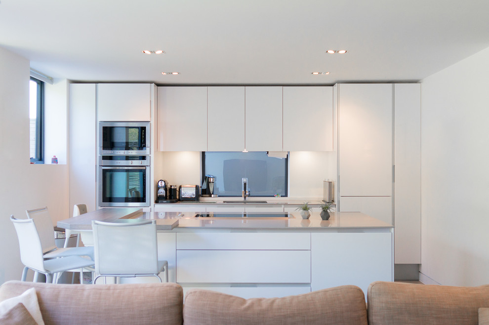 This is an example of a mid-sized contemporary single-wall open plan kitchen in Dorset with an integrated sink, flat-panel cabinets, white cabinets, quartz benchtops, white splashback, window splashback, panelled appliances, porcelain floors and with island.