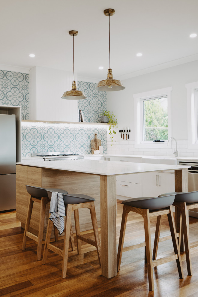 Photo of a kitchen in Hobart with a farmhouse sink, white cabinets, granite benchtops, multi-coloured splashback, cement tile splashback, stainless steel appliances, light hardwood floors and with island.