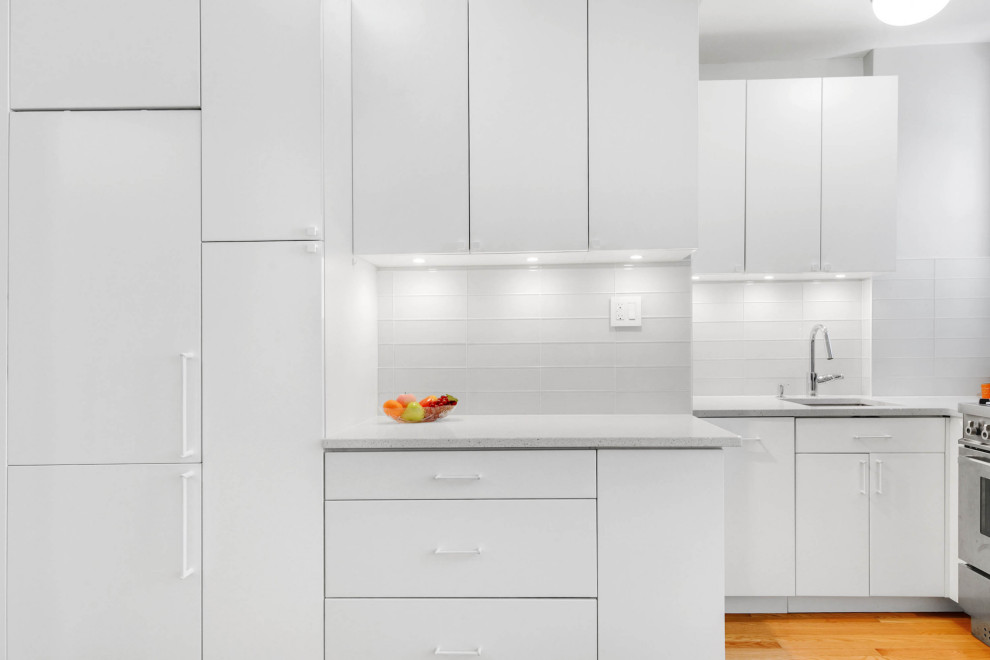 Inspiration for a small modern l-shaped kitchen pantry in New York with a submerged sink, flat-panel cabinets, white cabinets, engineered stone countertops, white splashback, glass tiled splashback, stainless steel appliances, medium hardwood flooring, orange floors and white worktops.
