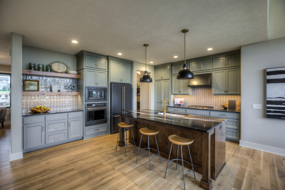 Photo of a traditional l-shaped kitchen in Omaha with an undermount sink, recessed-panel cabinets, green cabinets, grey splashback, black appliances, light hardwood floors, with island, beige floor and black benchtop.