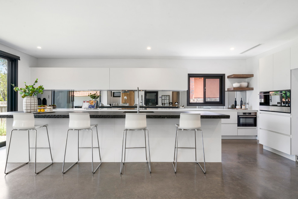 This is an example of a mid-sized contemporary galley open plan kitchen in Sydney with a double-bowl sink, granite benchtops, mirror splashback, stainless steel appliances, concrete floors, grey floor, flat-panel cabinets, white cabinets and with island.