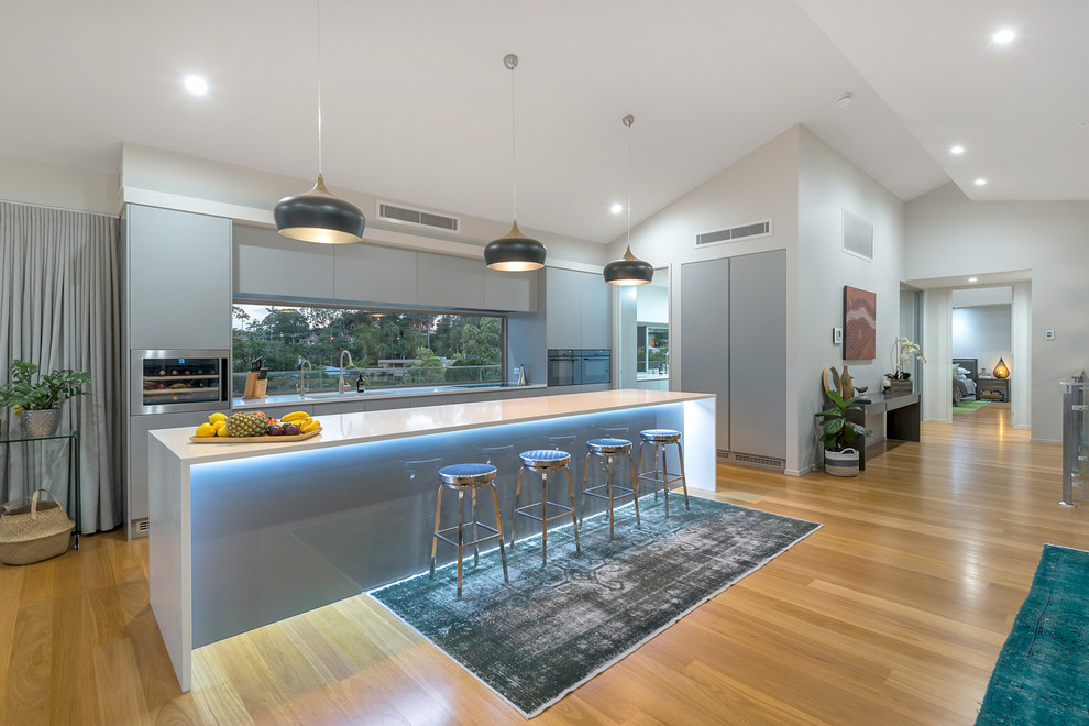 Mid-sized contemporary l-shaped eat-in kitchen in Brisbane with an undermount sink, grey cabinets, with island, brown floor, white benchtop, flat-panel cabinets, window splashback and medium hardwood floors.