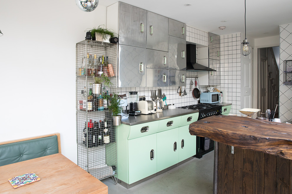 Large eclectic kitchen in London with green cabinets, stainless steel benchtops, concrete floors, with island, grey benchtop, flat-panel cabinets, white splashback, black appliances and grey floor.