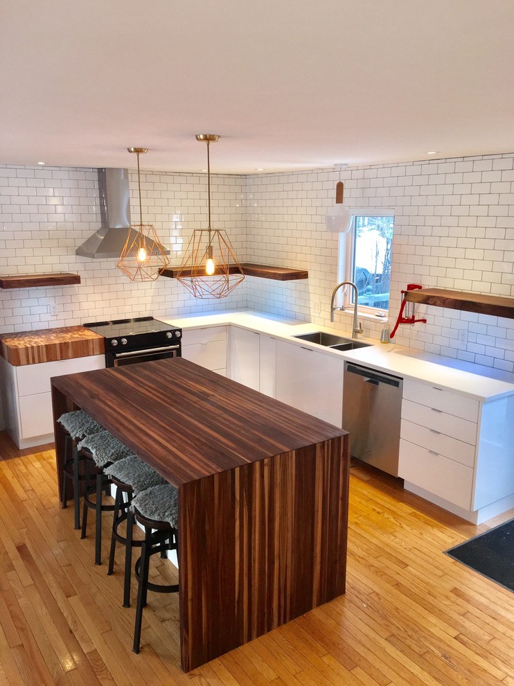 Mid-sized minimalist l-shaped medium tone wood floor eat-in kitchen photo with an undermount sink, flat-panel cabinets, white cabinets, solid surface countertops, white backsplash, subway tile backsplash, stainless steel appliances and an island