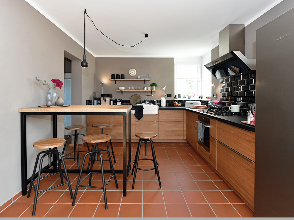 Inspiration for a mid-sized industrial l-shaped open plan kitchen in Frankfurt with a single-bowl sink, flat-panel cabinets, medium wood cabinets, wood benchtops, black splashback, subway tile splashback, stainless steel appliances, terra-cotta floors, a peninsula, red floor and black benchtop.