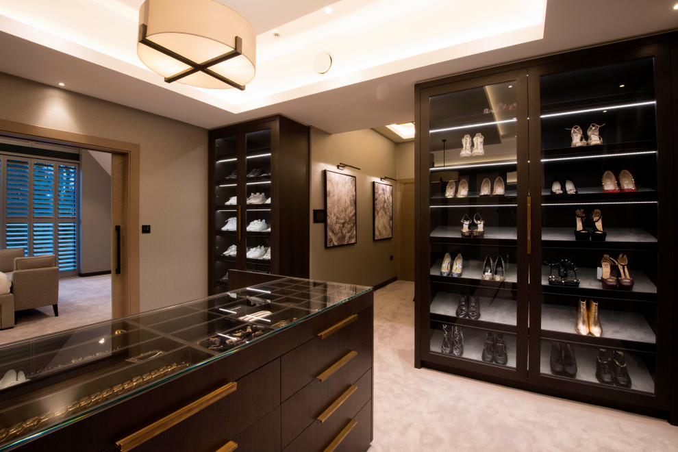 This is an example of an expansive contemporary gender neutral dressing room in Cheshire with glass-front cabinets, dark wood cabinets, carpet, beige floors, a coffered ceiling and feature lighting.