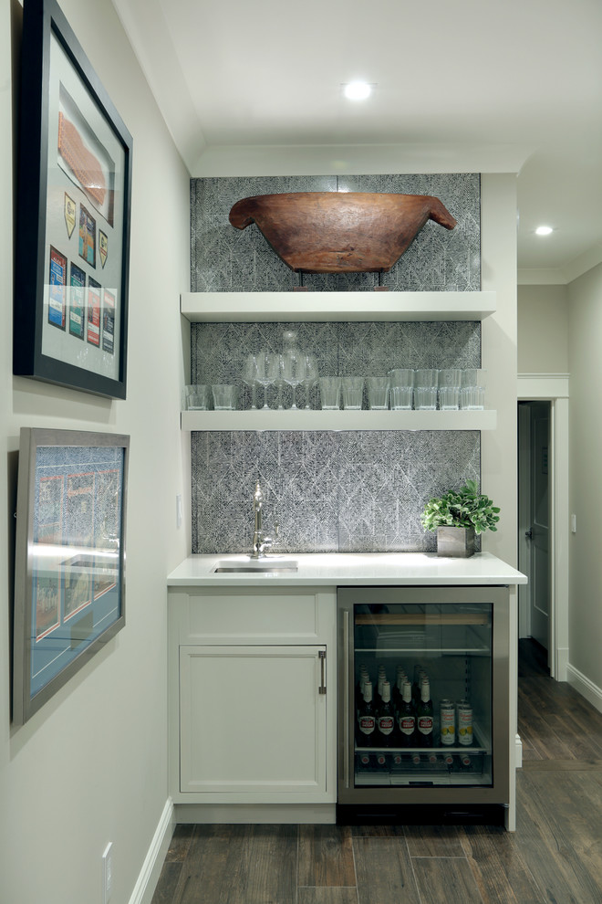 Design ideas for a mid-sized country single-wall wet bar in Grand Rapids with an undermount sink, recessed-panel cabinets, white cabinets, quartz benchtops, grey splashback, ceramic splashback, medium hardwood floors, brown floor and white benchtop.