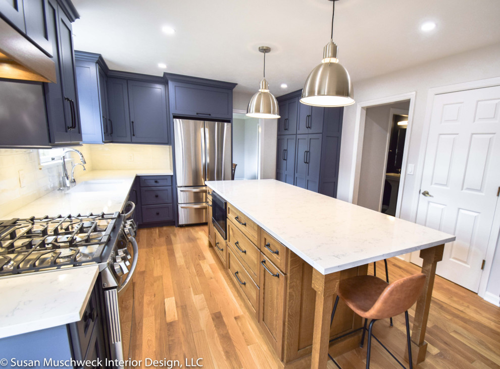 Mid-sized transitional l-shaped light wood floor enclosed kitchen photo with an undermount sink, shaker cabinets, blue cabinets, quartz countertops, white backsplash, ceramic backsplash, stainless steel appliances, an island and white countertops
